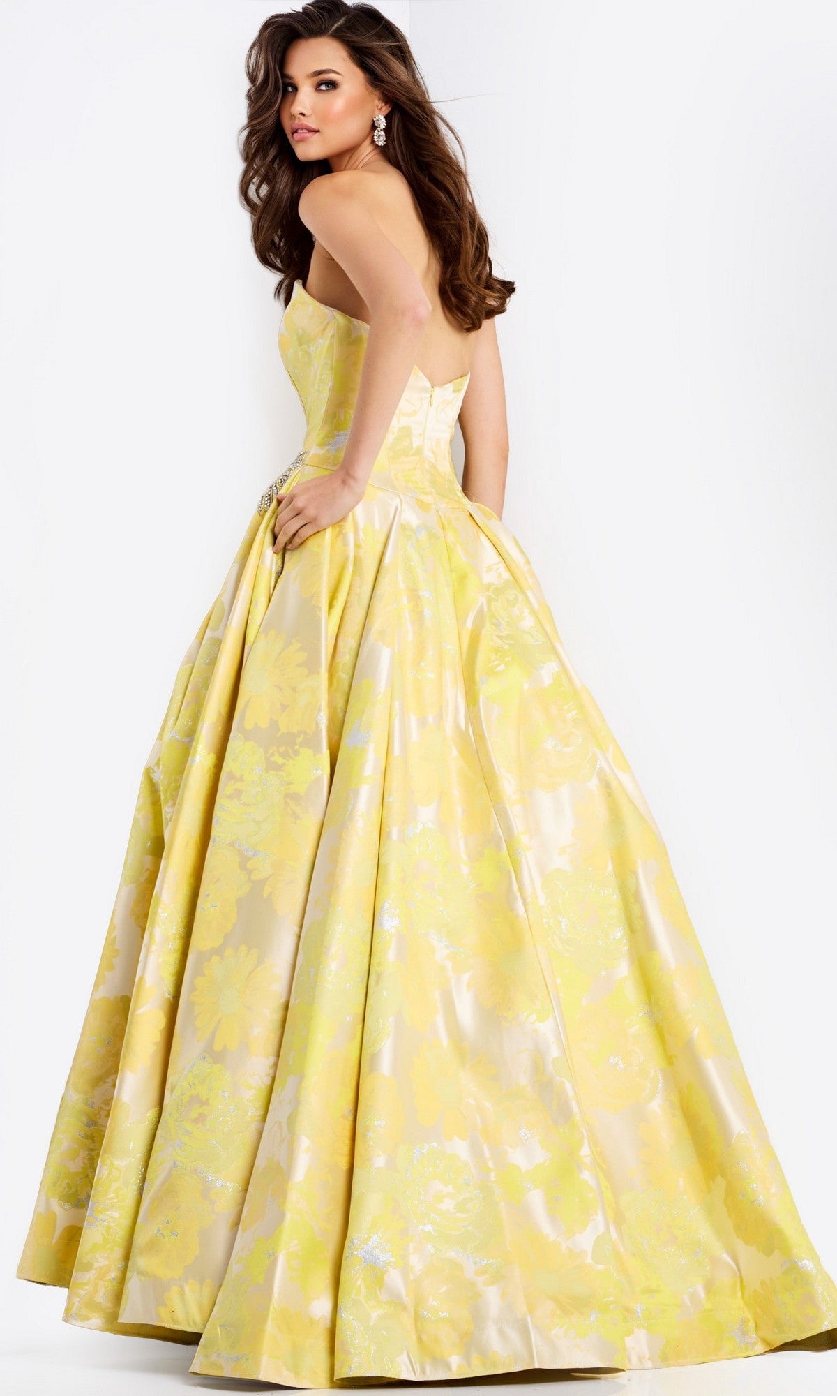 A woman poses with her back turned, looking over her shoulder in the Strapless Long Yellow Prom Ball Gown: Jovani 49502, featuring a full skirt and floral metallic print against a plain background.